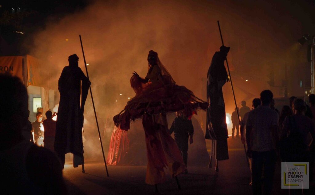 Performers on stilts walking through smoke on street at night
