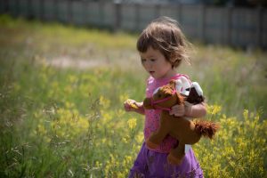 Young girl walking towards camera in field of grass and yellow flowers holding two toy ponies