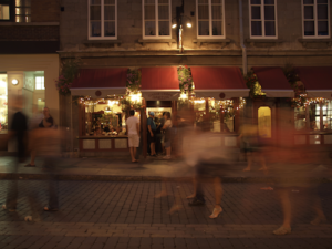 Ghosting of people walking along European style street with lit up shops and customers in background at night