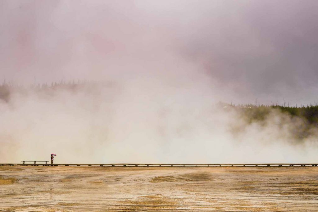Person standing on edge of boardwalk overlooking geysers with steam rising